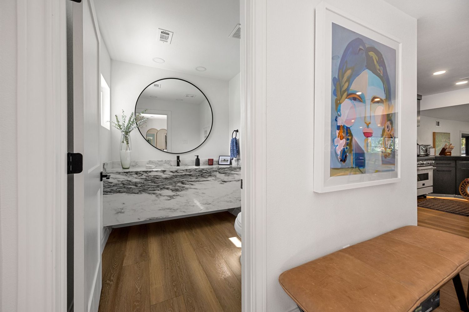 Modern bathroom with marble vanity, round black-framed mirror, and hardwood floors viewed through a doorway with colorful wall art.