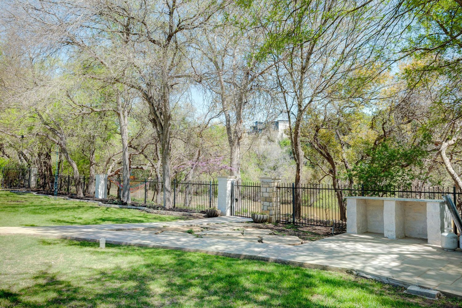 Tree-lined property with ornate iron gate, paved driveway, and white garage structure in a lush green landscape.