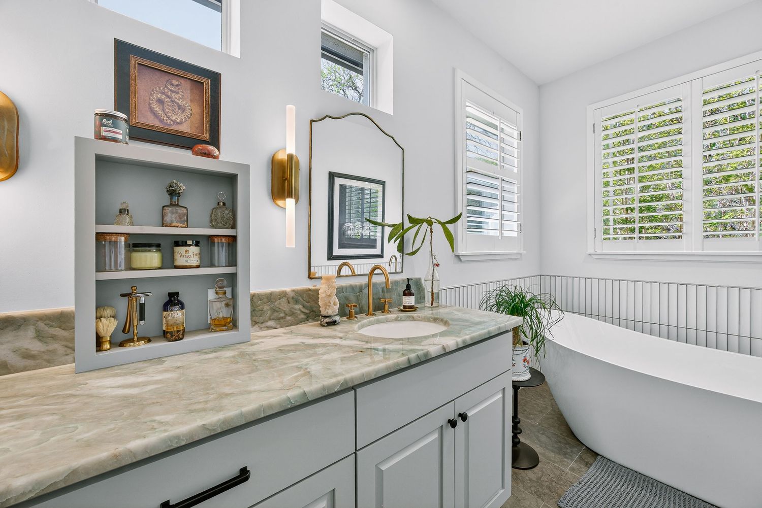 Elegant bathroom with gray vanity, marble countertop, built-in shelves, gold mirror, and freestanding soaking tub.