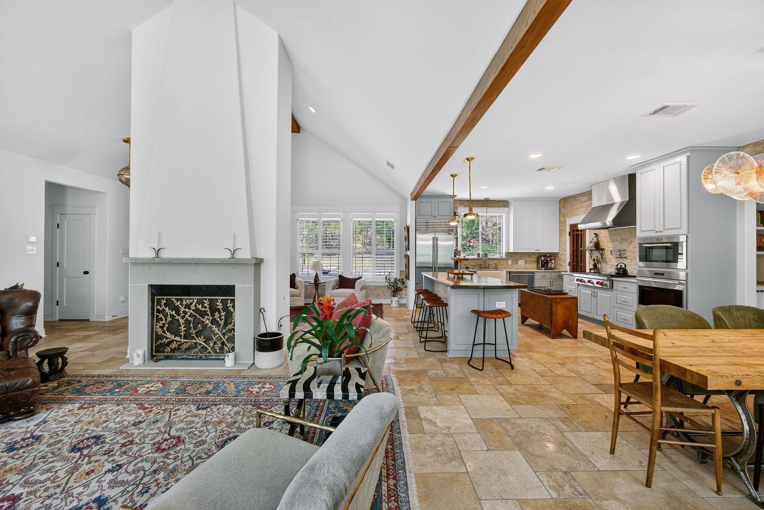 Open-concept living room with fireplace flows into modern kitchen featuring island seating, stone tile floors, and exposed wood beams.