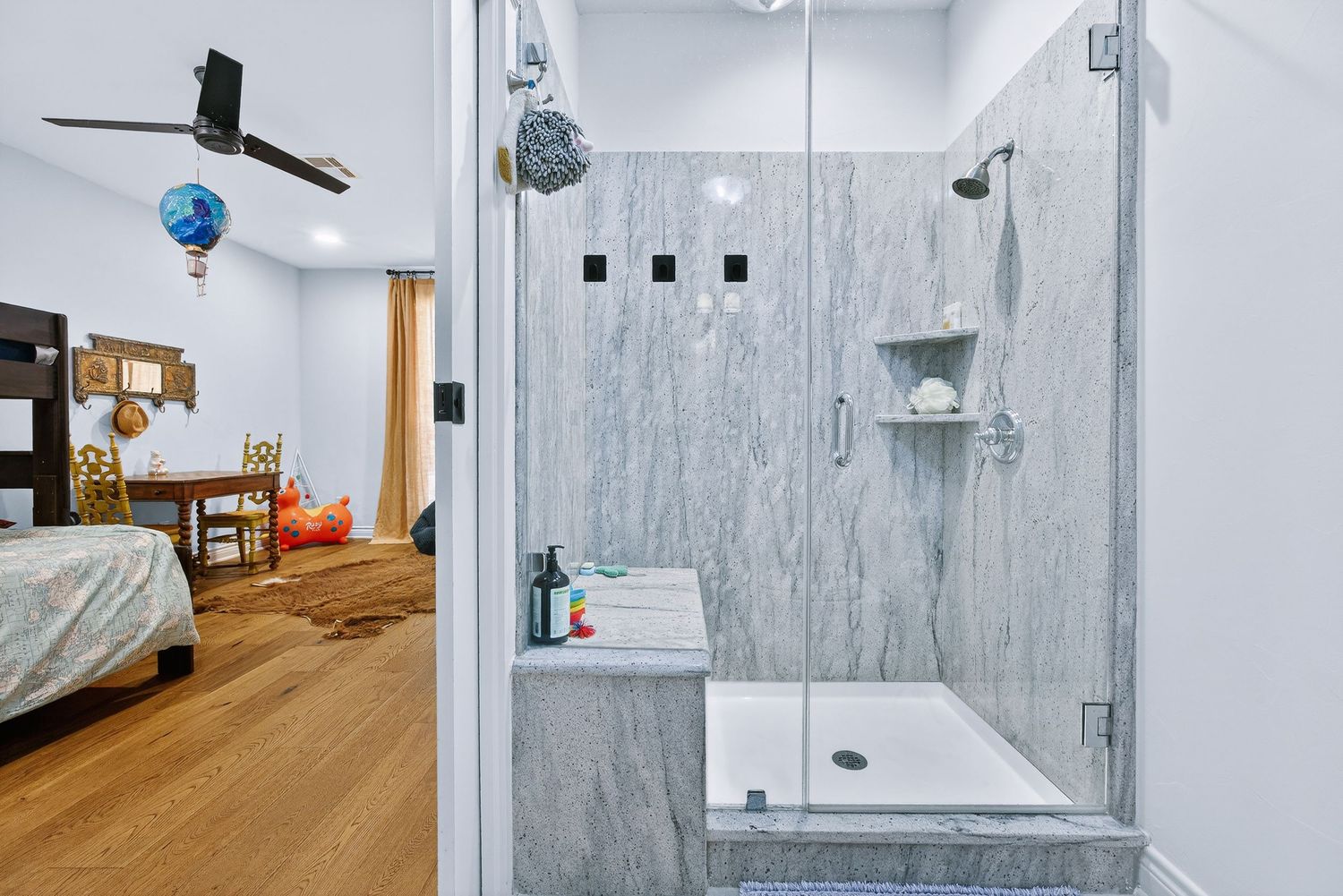 Modern bathroom with marble walk-in shower and glass door, open to a hardwood floor bedroom with ceiling fan.