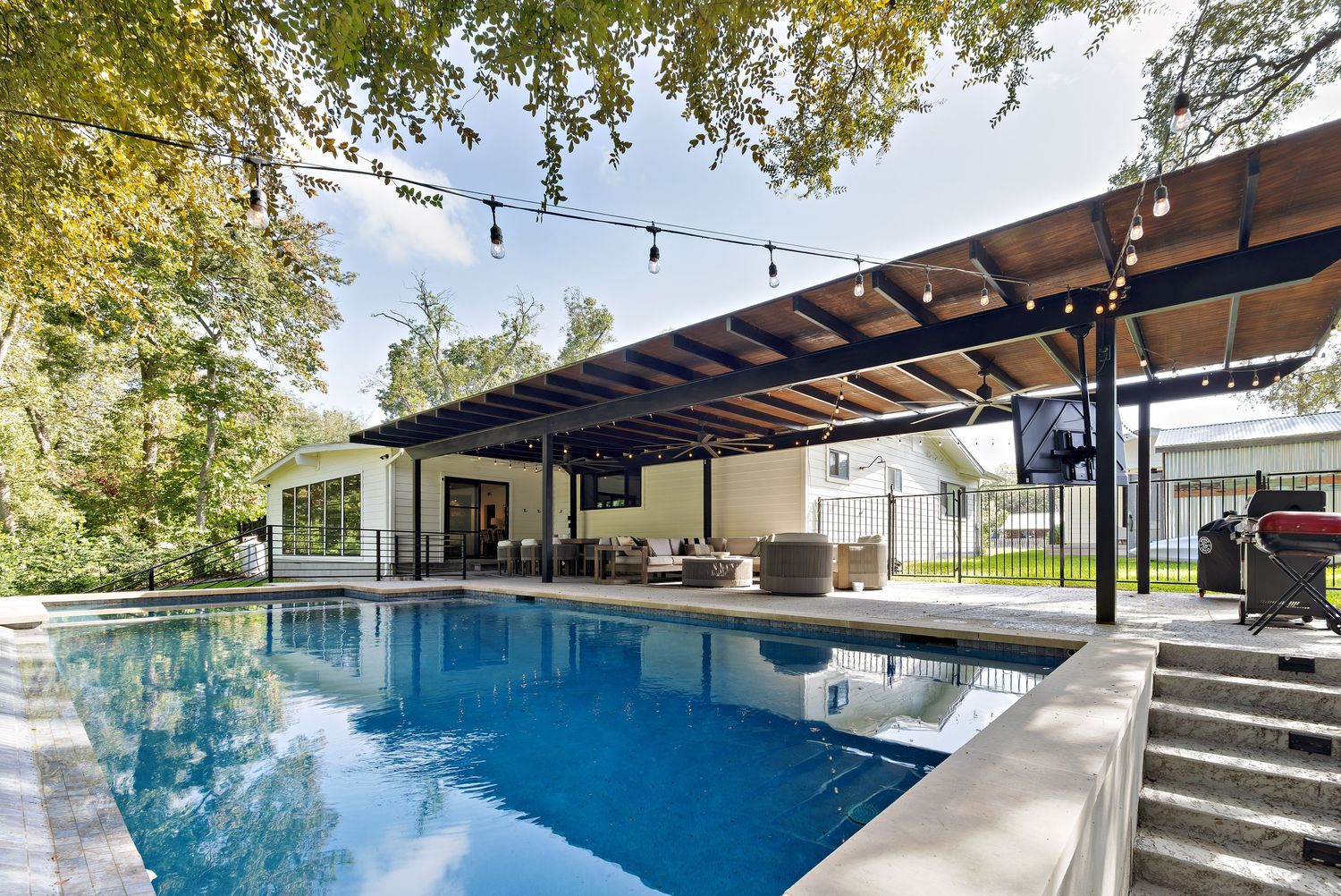 Outdoor pool with pergola, string lights, lounge seating, and lush tree canopy in backyard.
