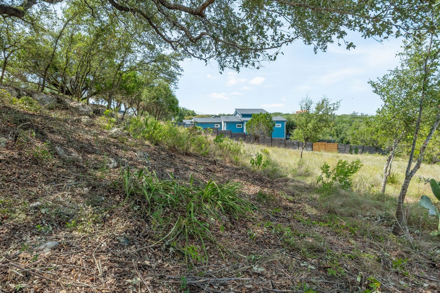 A sloped hillside with dry grass and trees overlooks a blue building and lush greenery under a partly cloudy sky.