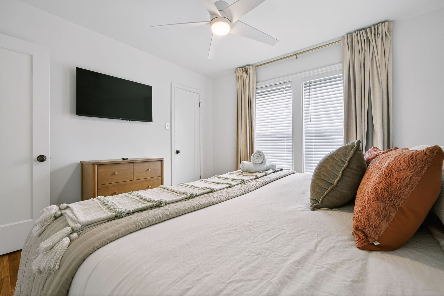 Modern bedroom interior with ceiling fan, white walls, wooden furniture, and orange accent pillows.