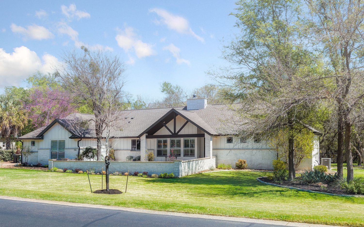 Single-story white ranch home with Tudor-style gable, lush green lawn, and mature trees under a bright blue sky.