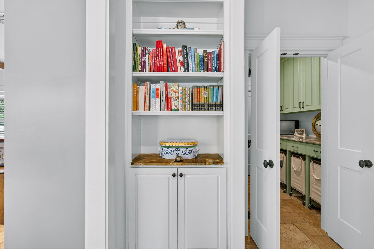 White built-in bookshelf with colorful books and cabinet doors, adjacent to a kitchen with green cabinets.