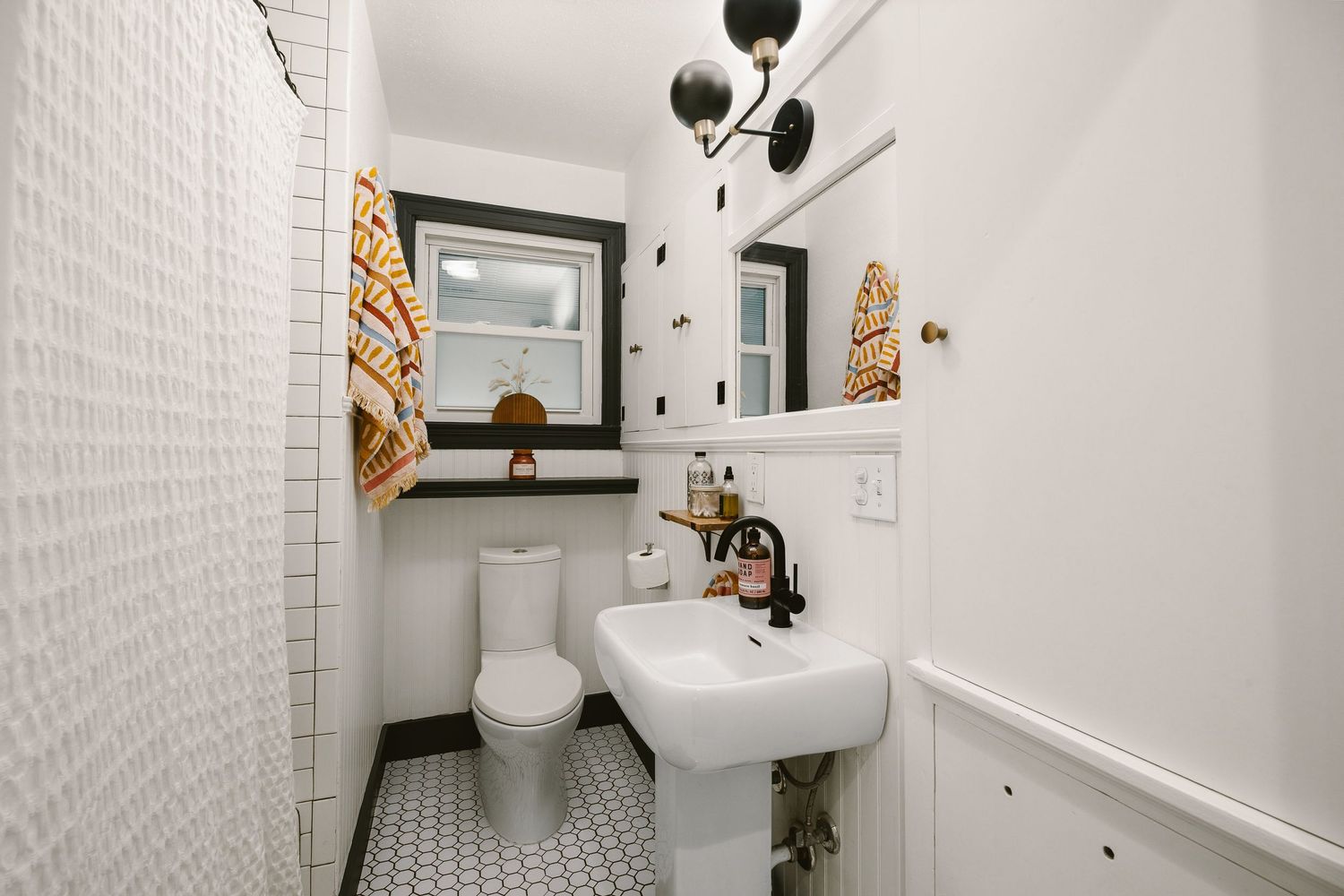 White bathroom with pedestal sink, black-framed mirror, patterned tile floor, and decorative wall sconce lighting.