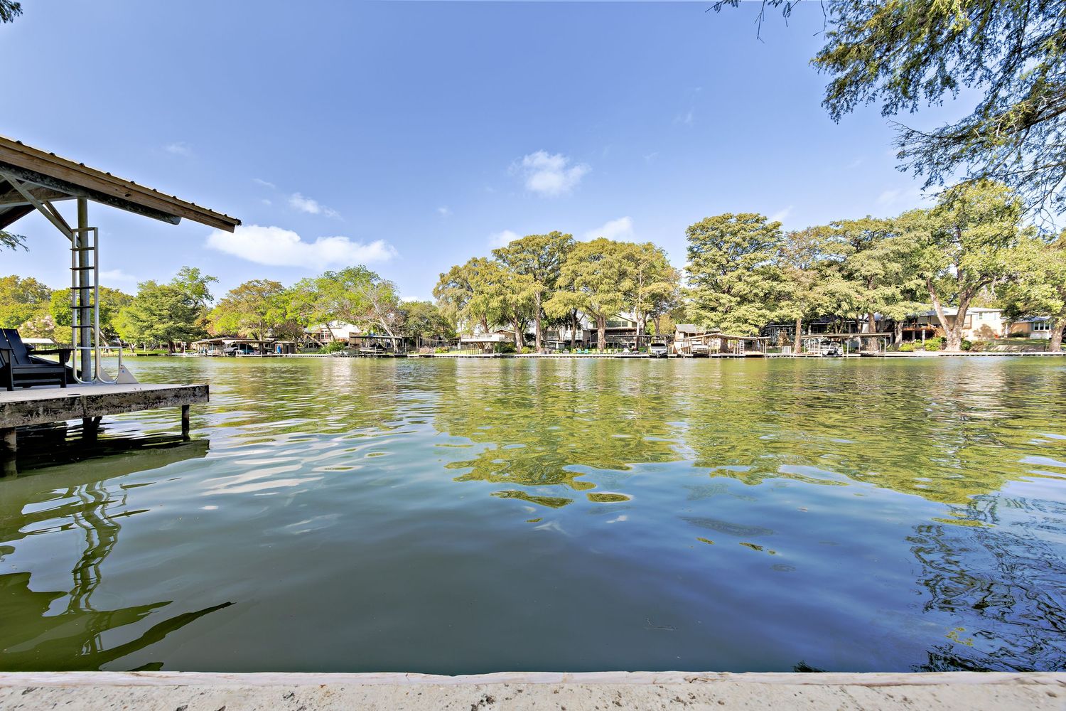 Tranquil lakefront view with calm green water, mature trees, a wooden dock, and neighboring homes along the shoreline.