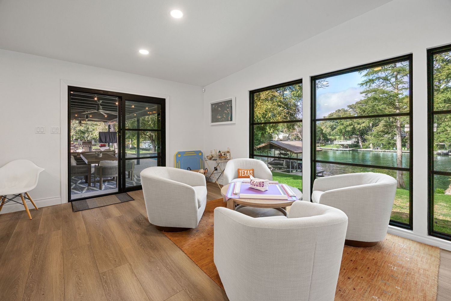 Bright sunlit sitting room with white barrel chairs, wood floors, and large black-framed windows overlooking a serene waterfront.