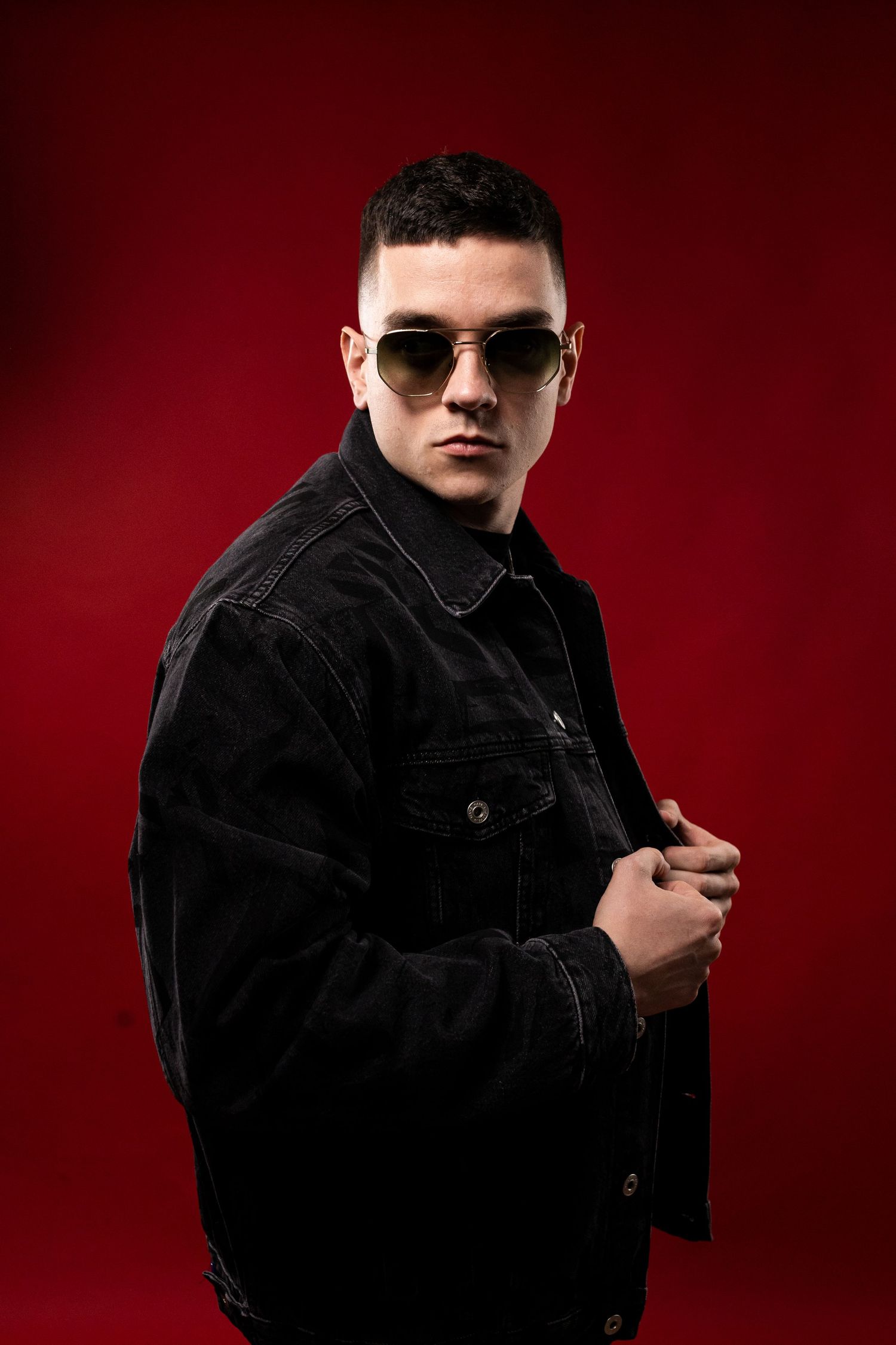 Young man in black denim jacket wearing gold aviator sunglasses posing on red background.