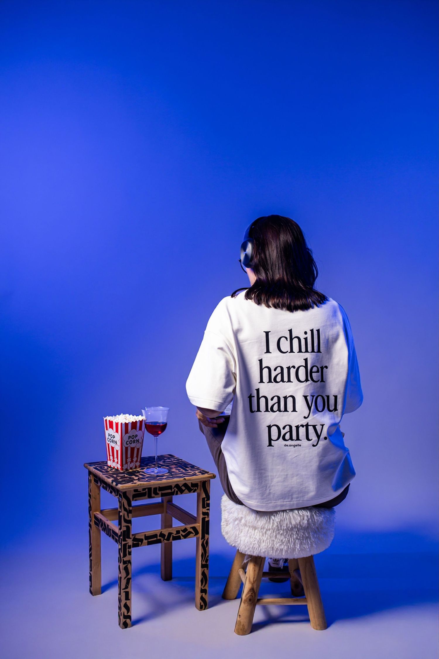 Person sitting on stool wearing white oversized tee reading 'I chill harder than you party' with popcorn and wine nearby.