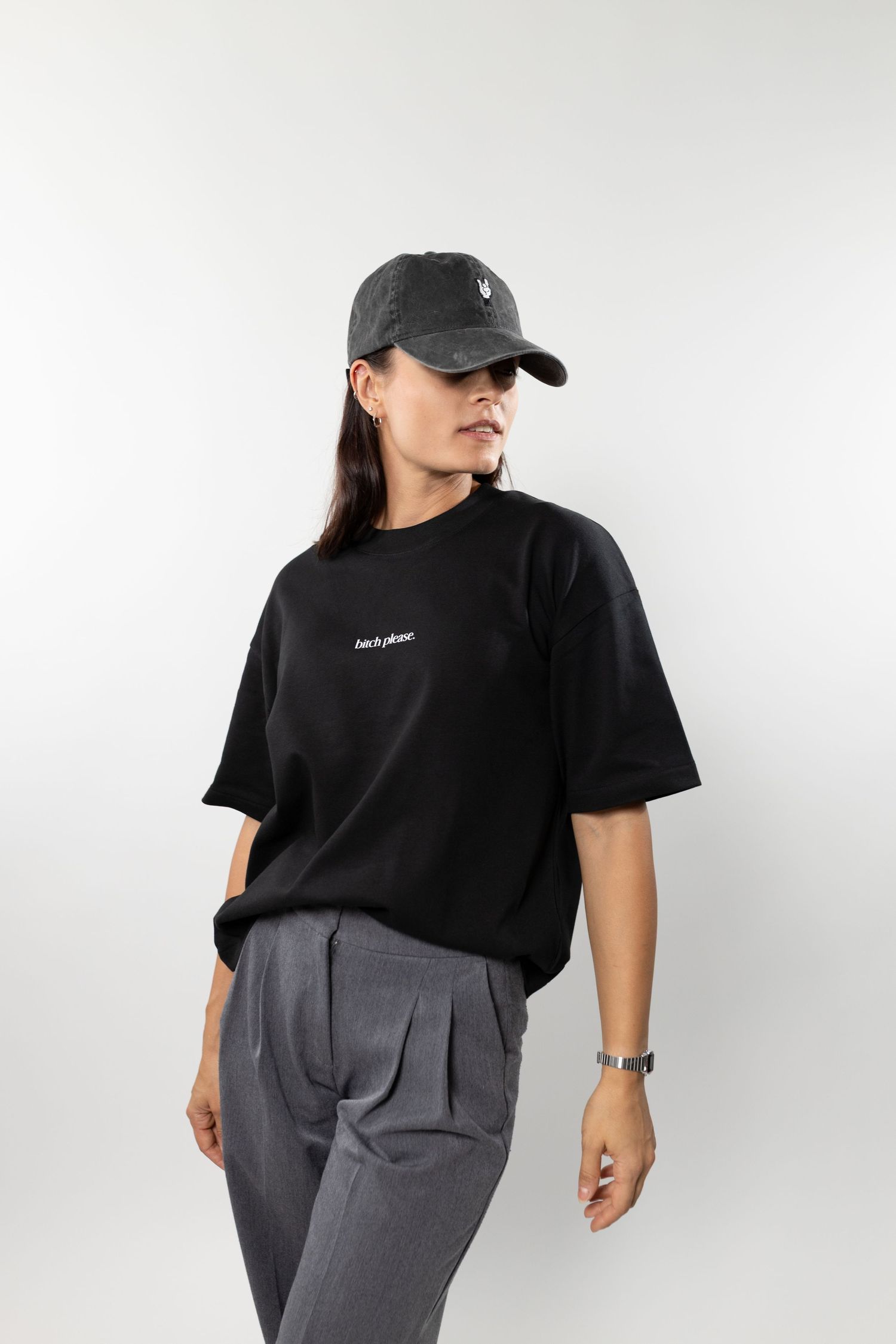 Woman wearing black oversized t-shirt, grey cap, and grey trousers, posing against white background.