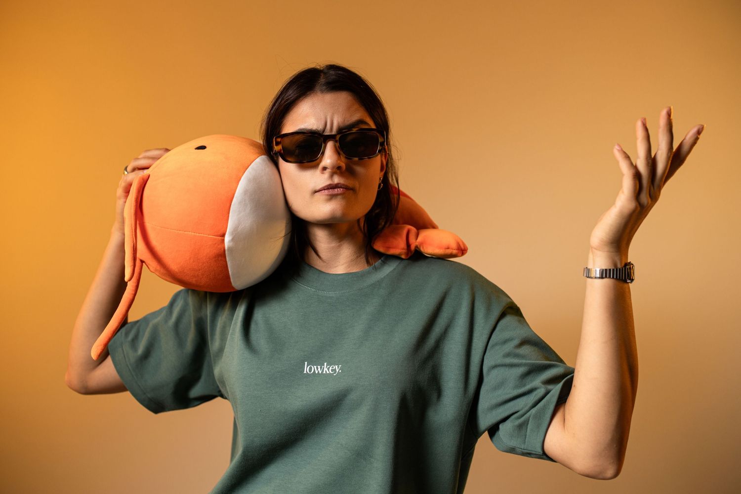 Woman in green oversized tee and sunglasses holding an orange ball on her shoulder against a beige background.