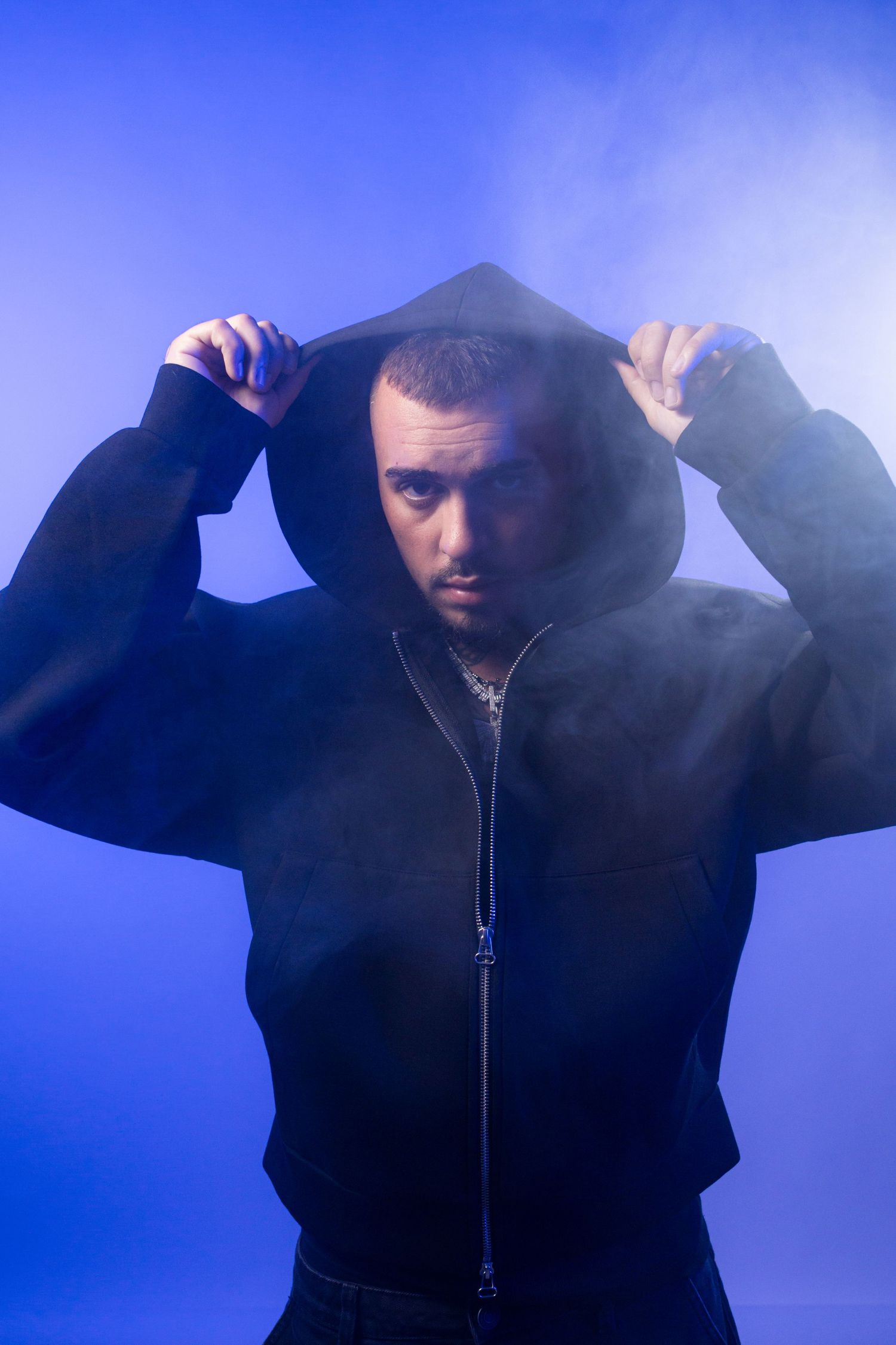 Man in black hoodie pulling up hood against blue smoky background.