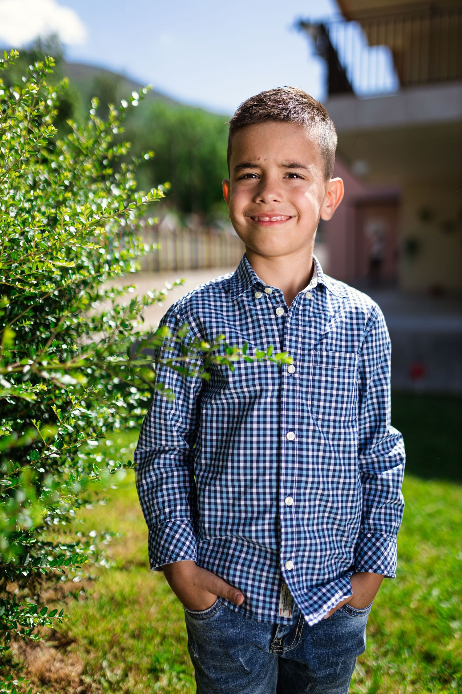 Smiling boy in blue plaid shirt standing outdoors with hands in pockets near green bushes on a sunny day.