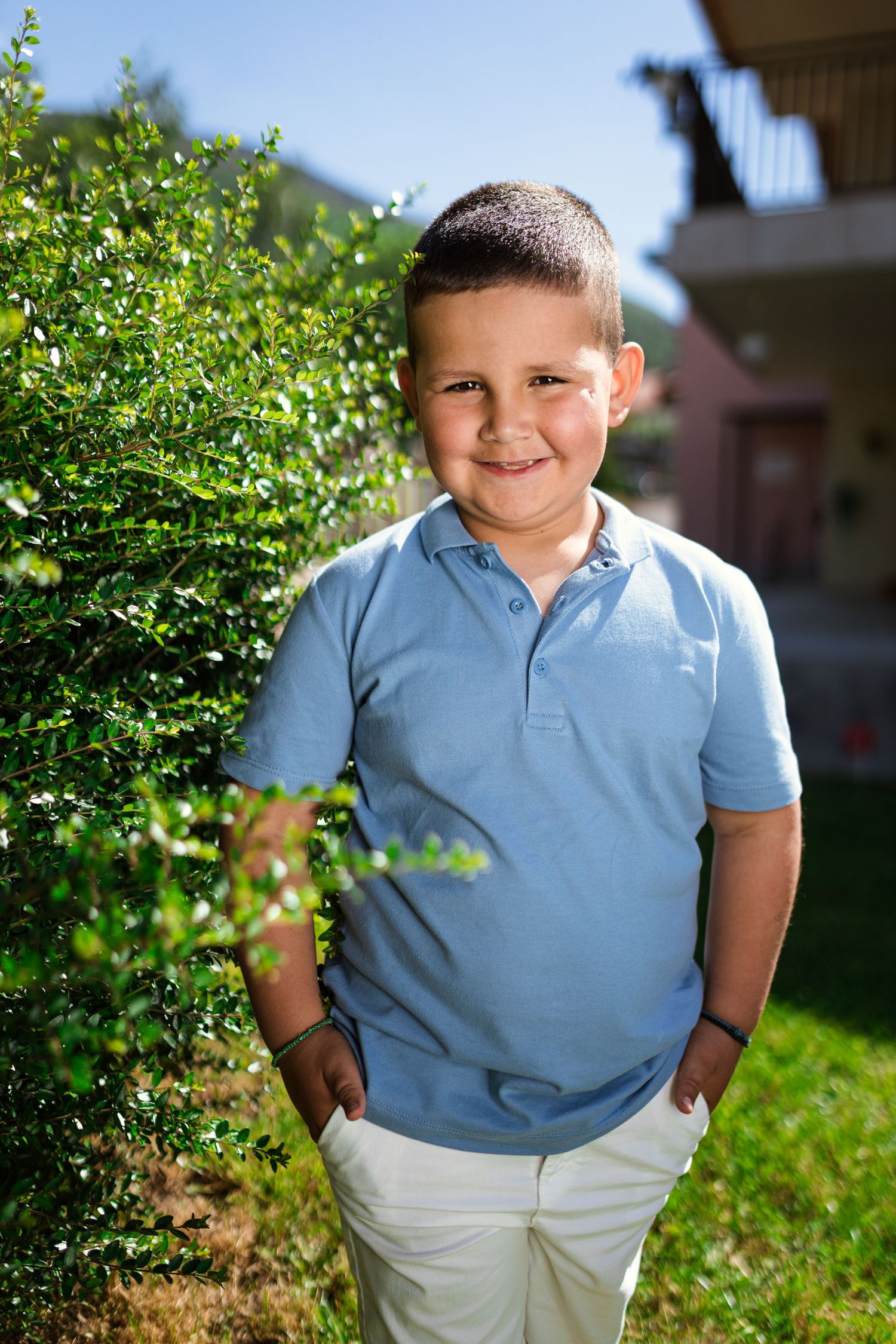 Smiling young boy in blue polo shirt and white pants standing outdoors near green bushes on a sunny day.