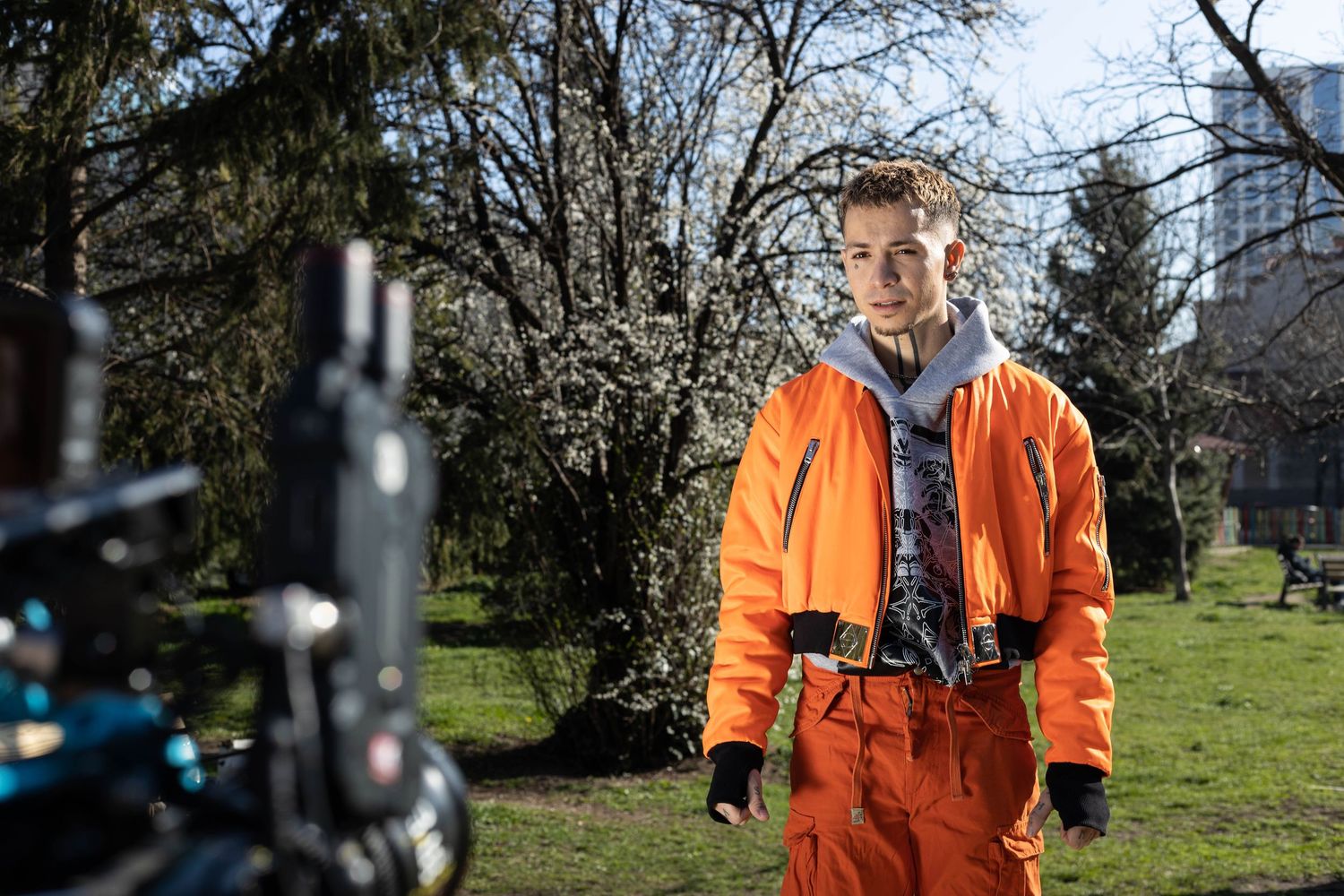 Young person in orange jumpsuit stands outdoors near blooming trees with camera equipment visible in foreground.
