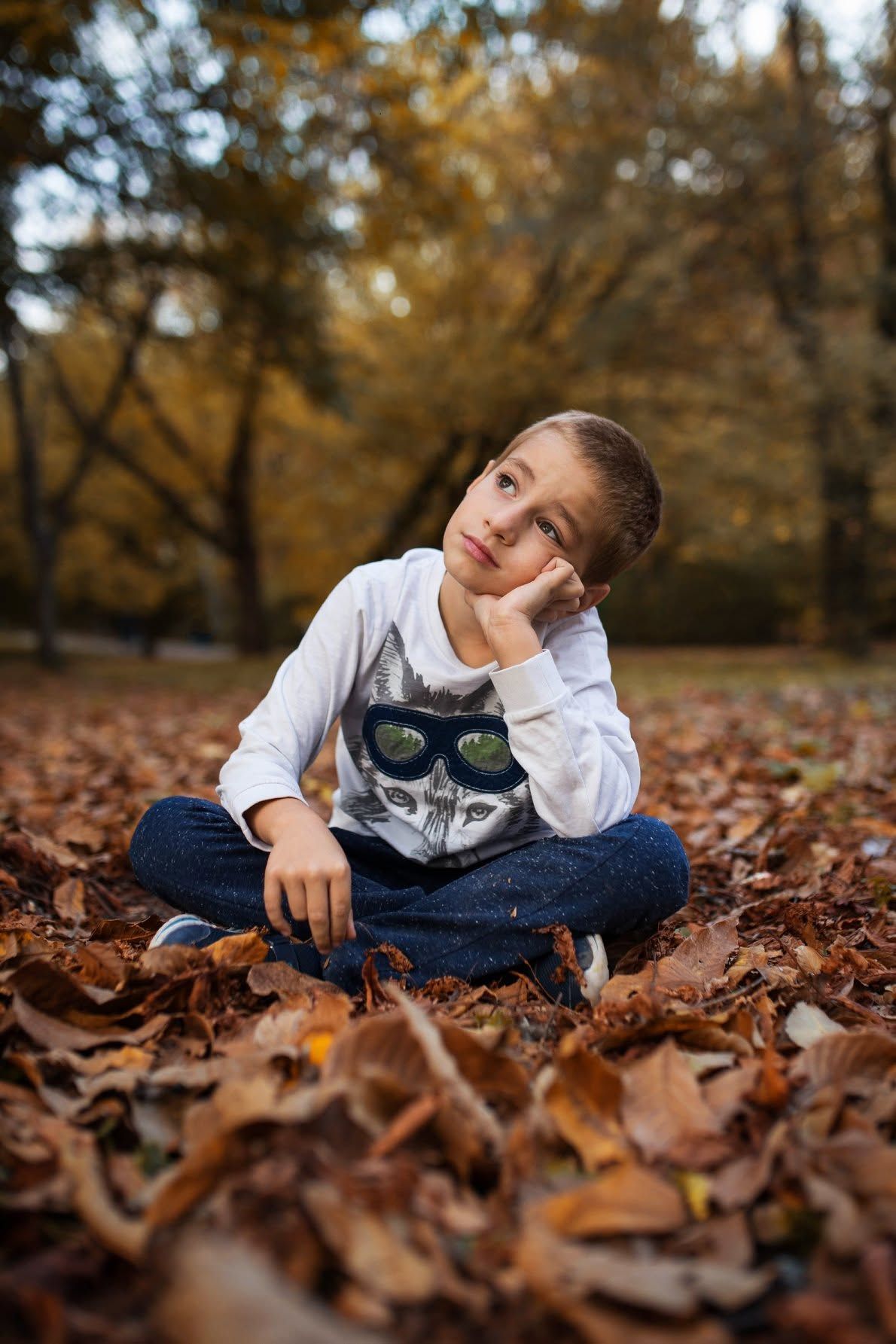 A thoughtful boy sits cross-legged on autumn leaves in a park, wearing a cat graphic sweatshirt and jeans.