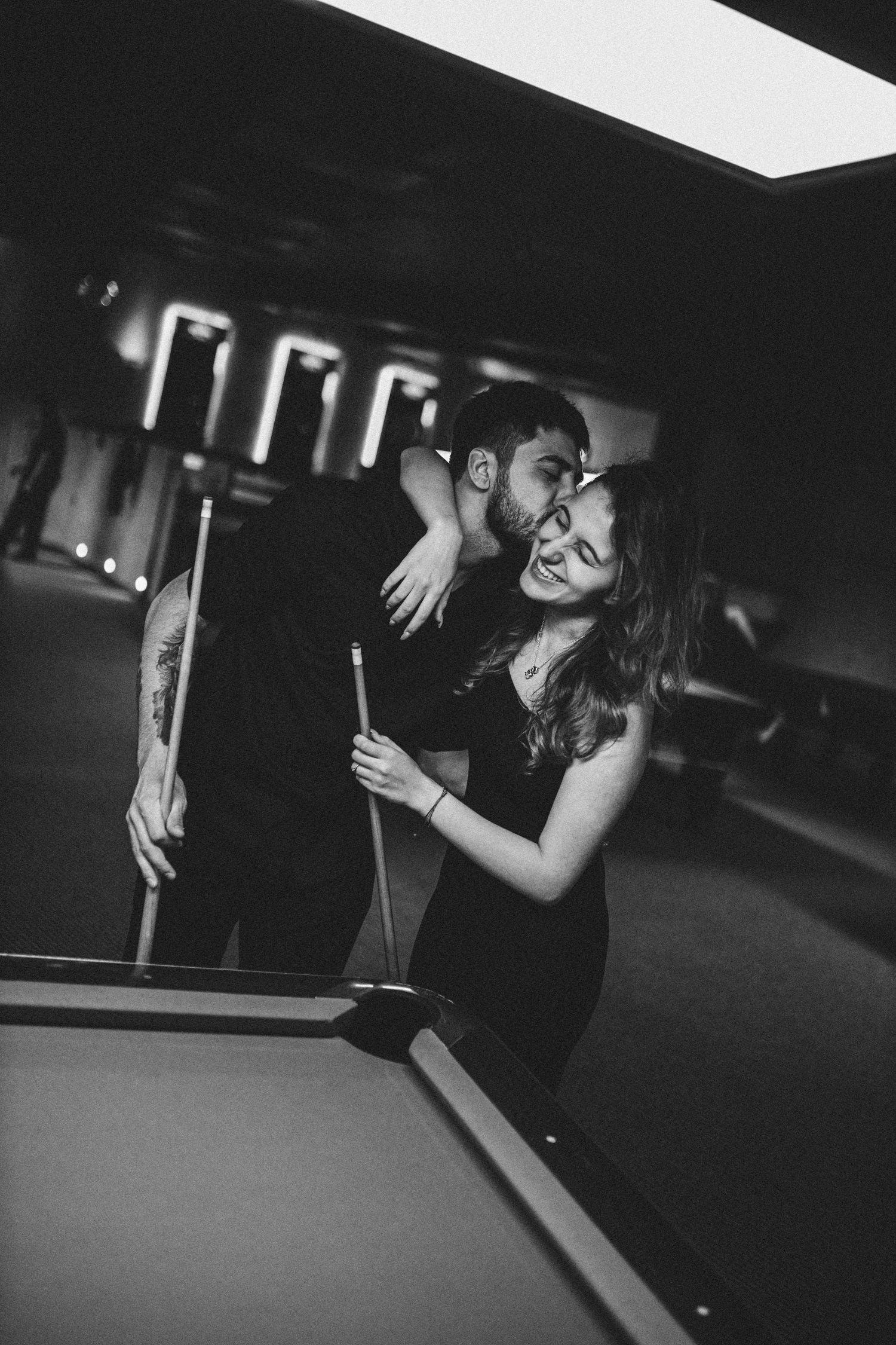 Black and white photo of a couple sharing a tender kiss beside a pool table in a billiard hall.