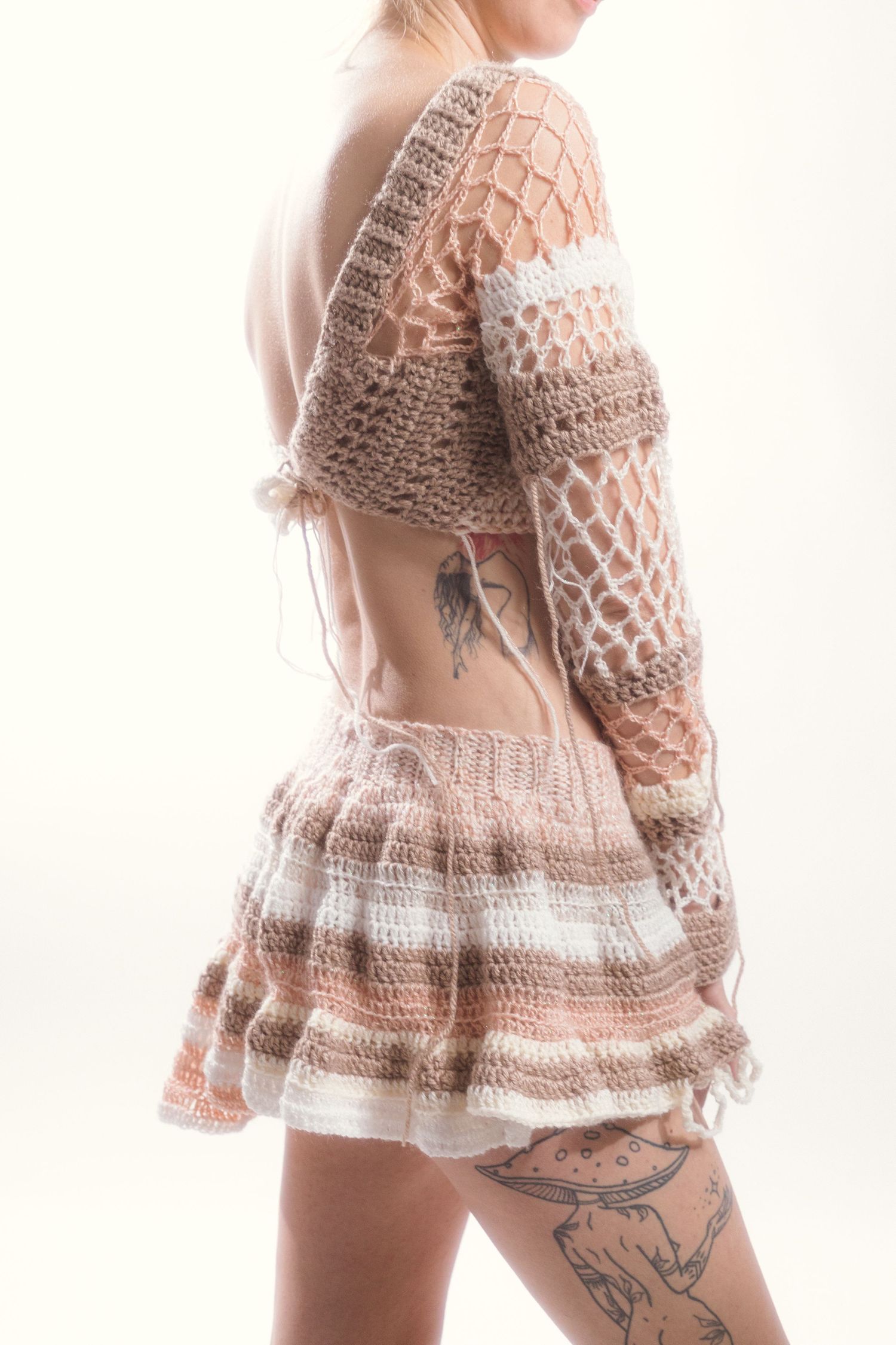 Back view of model wearing open-back crochet beige mini dress with thigh tattoo.