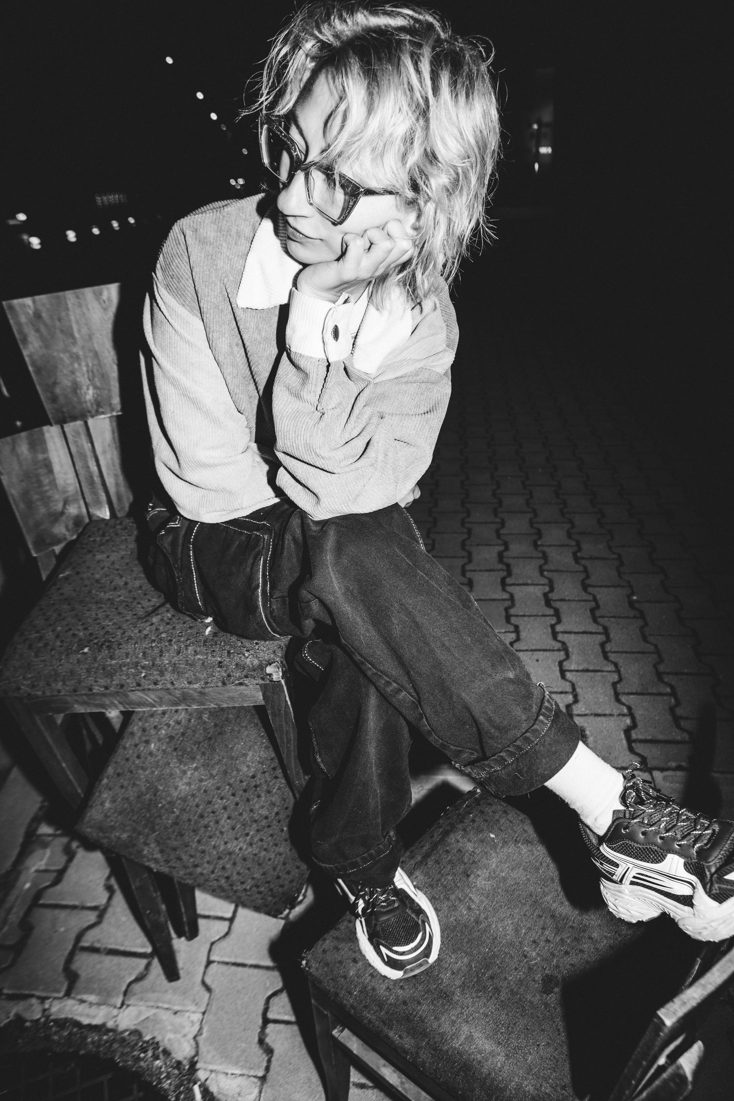 Black and white photo of a blonde woman with glasses sitting on a chair outdoors at night.