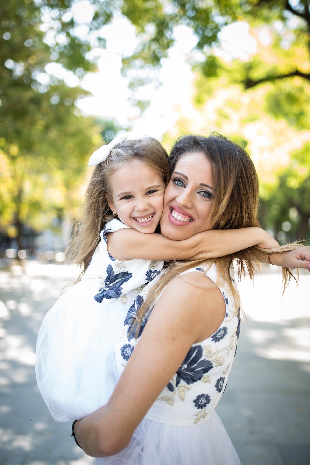 A smiling woman holds a happy young girl in a warm embrace outdoors in a sunny park.