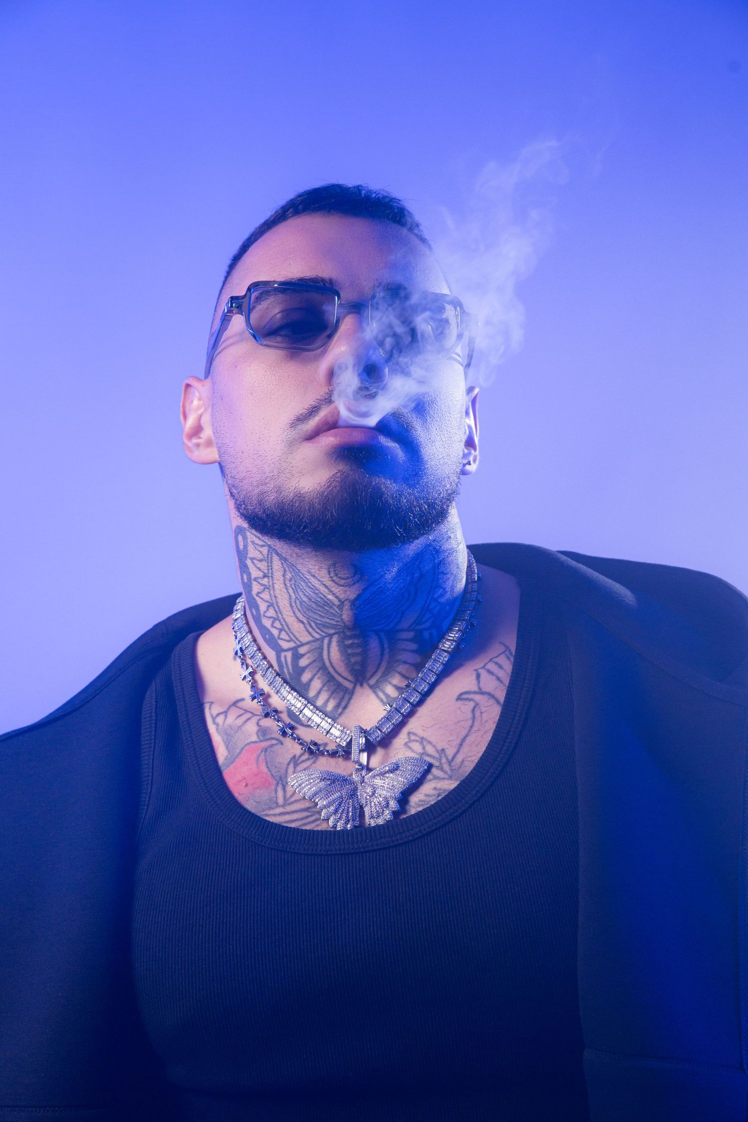 Tattooed man with sunglasses exhaling smoke against a blue background, wearing a black jacket and chain necklace.