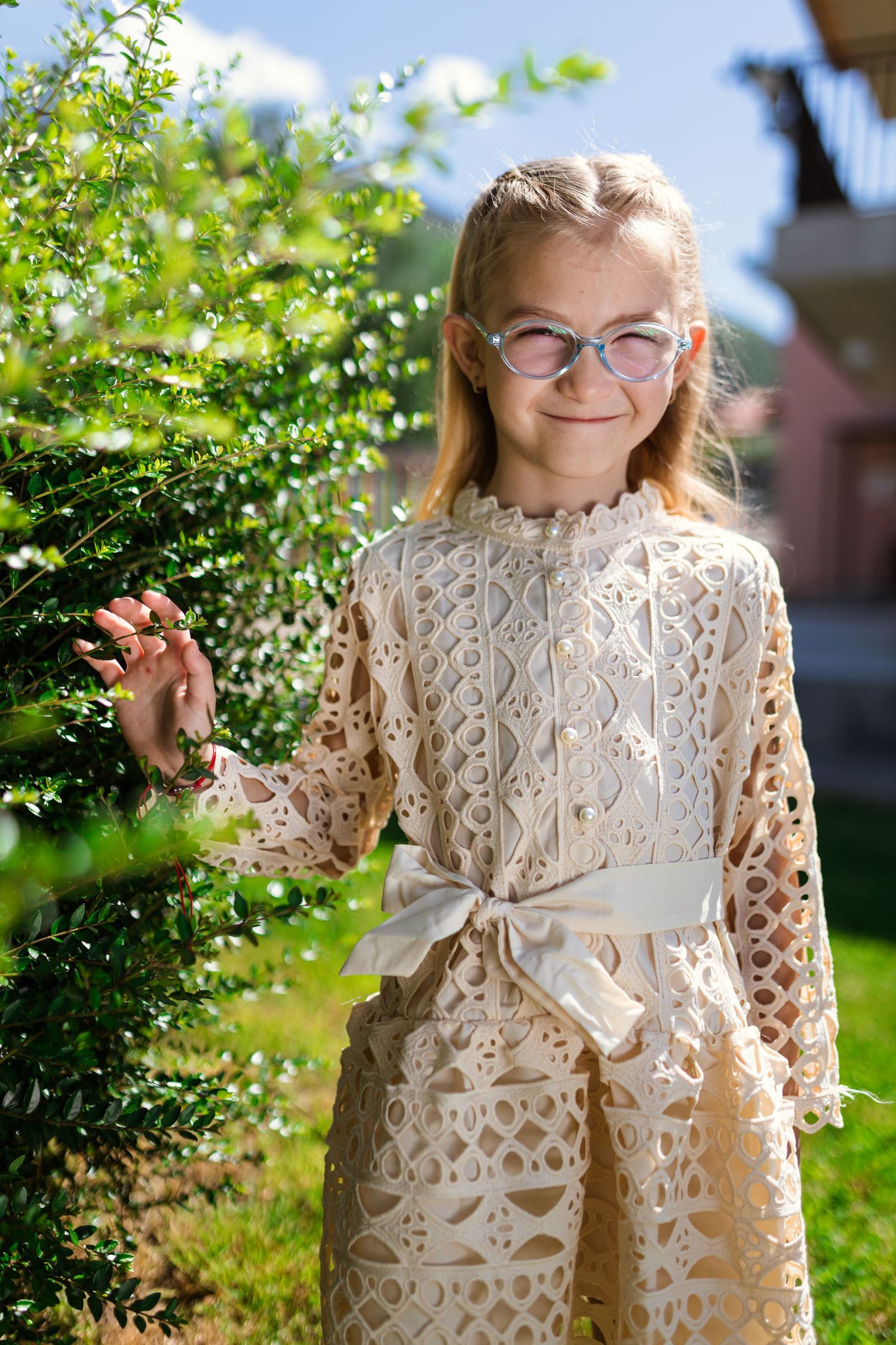 Young blonde girl in cream lace dress and glasses smiling outdoors near green bushes on a sunny day.