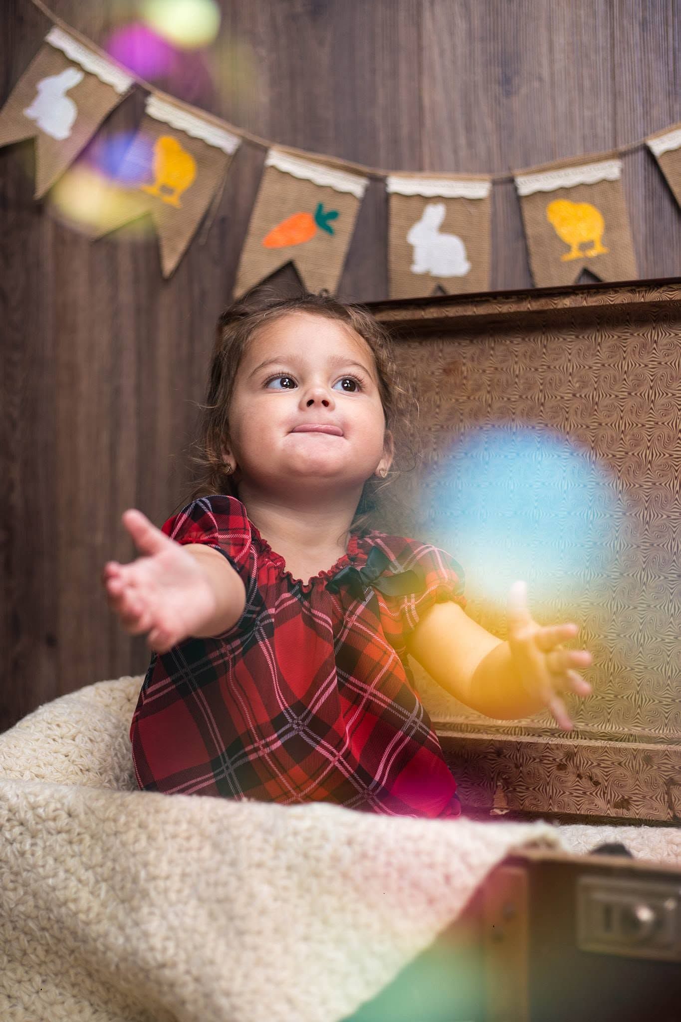 A toddler girl in a plaid dress sits in a vintage suitcase, reaching toward colorful lights with Easter bunting decorations behind her.