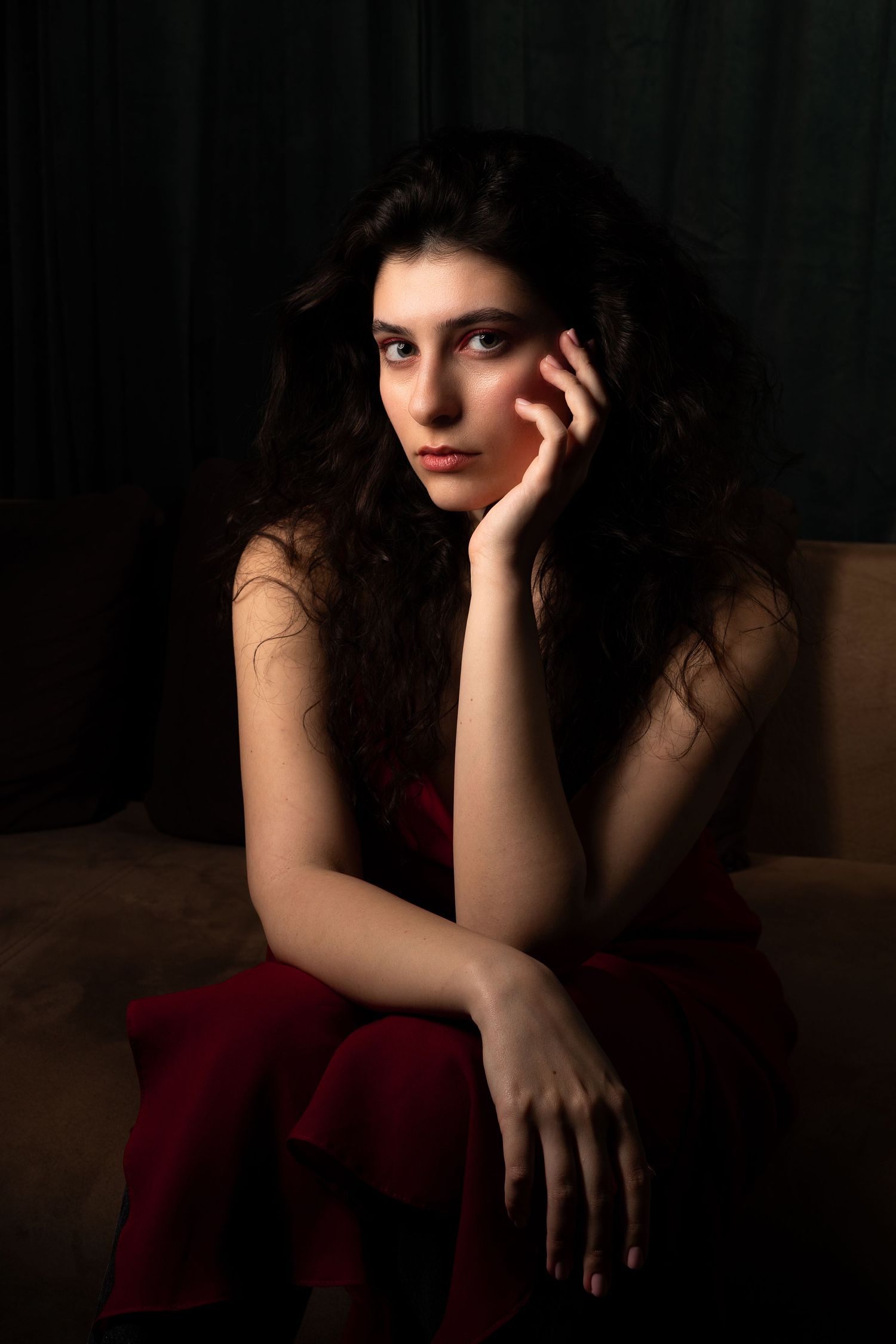 Young woman in red dress sitting pensively with hand on cheek in dramatic dark studio lighting.
