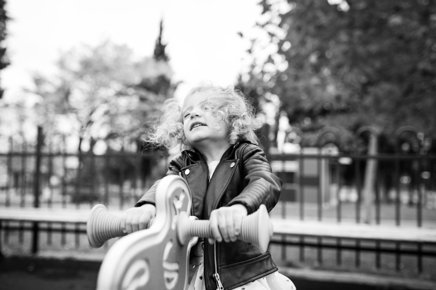 Curly-haired child in leather jacket riding playground toy with joy in black and white photo.