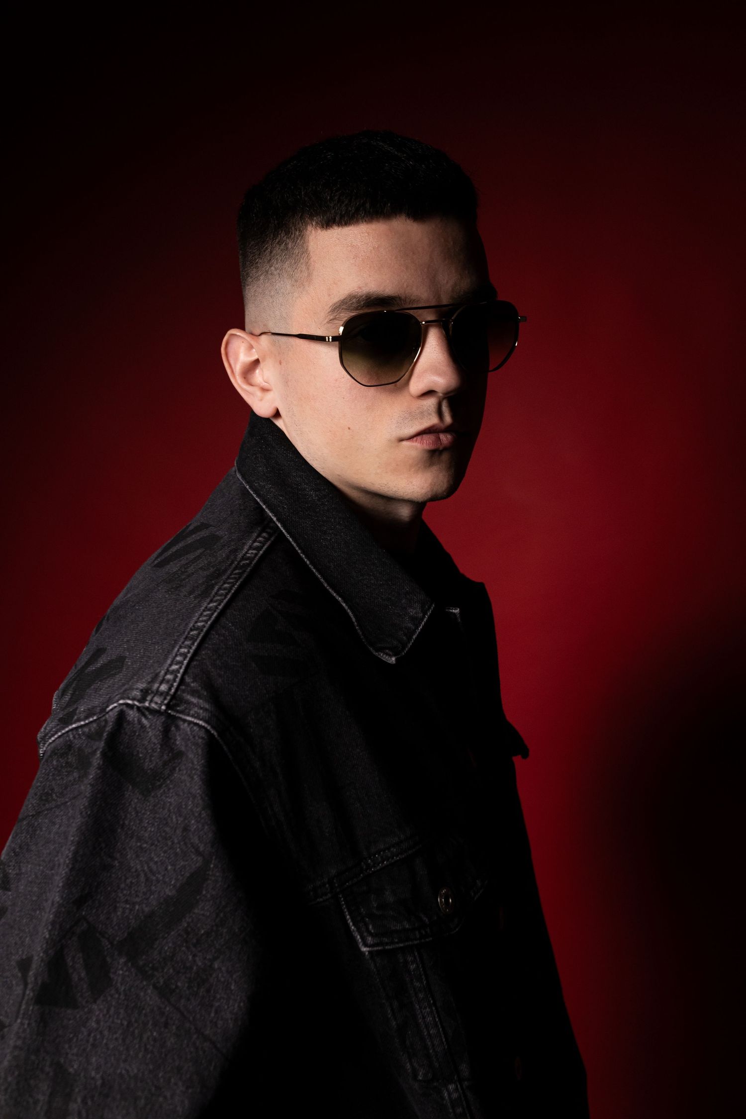 Stylish young man in black denim jacket wearing aviator sunglasses against red background.