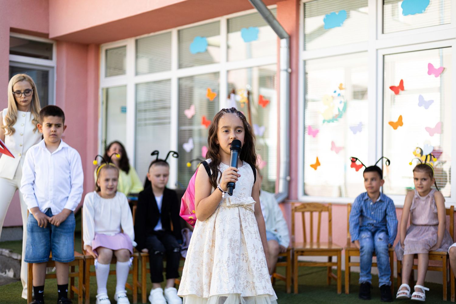A young girl in a white dress speaks into a microphone at an outdoor school event with children seated behind her.