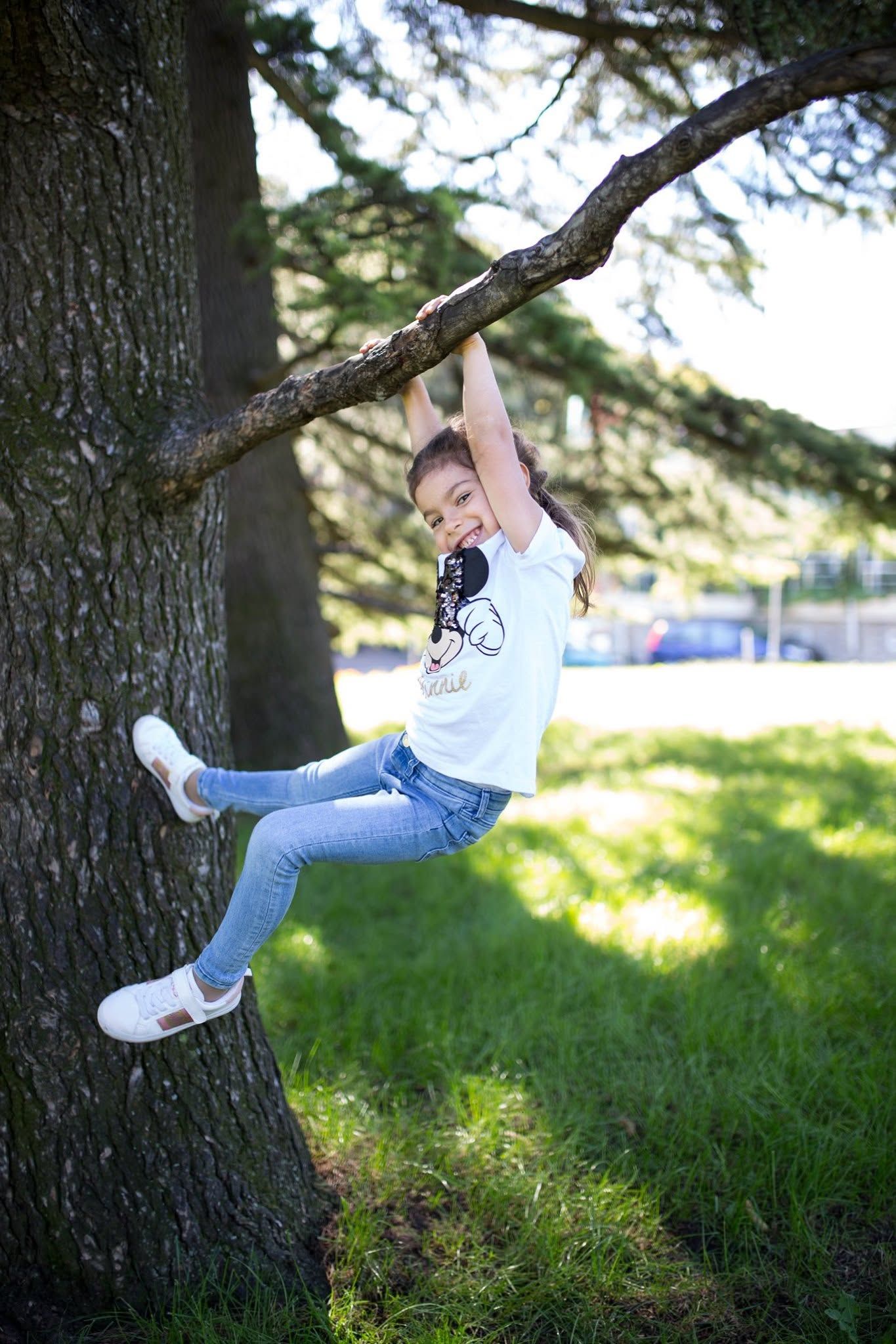 Young girl in jeans and white tee smiling while hanging from a tree branch outdoors in a sunny park.