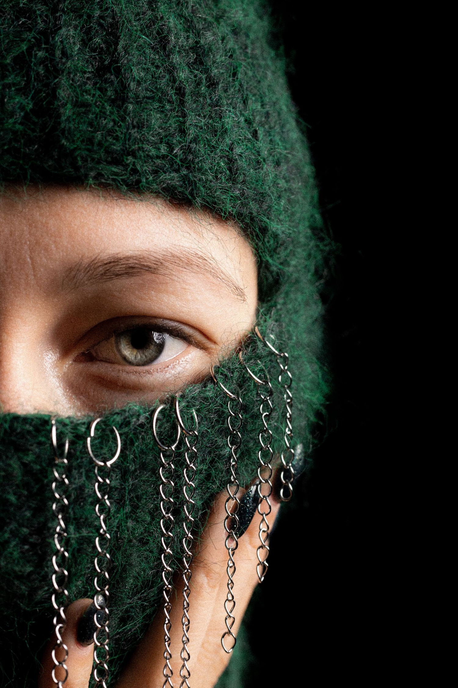 Close-up portrait of a person wearing a green knit balaclava adorned with silver chains and rings.