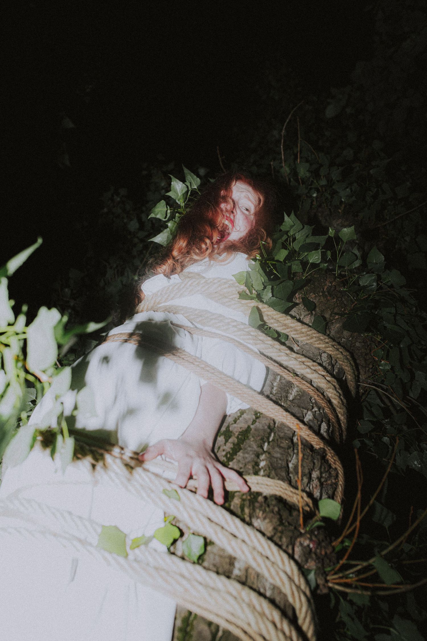 Red-haired woman in white dress wrapped in rope against moss-covered tree in dark outdoor setting.