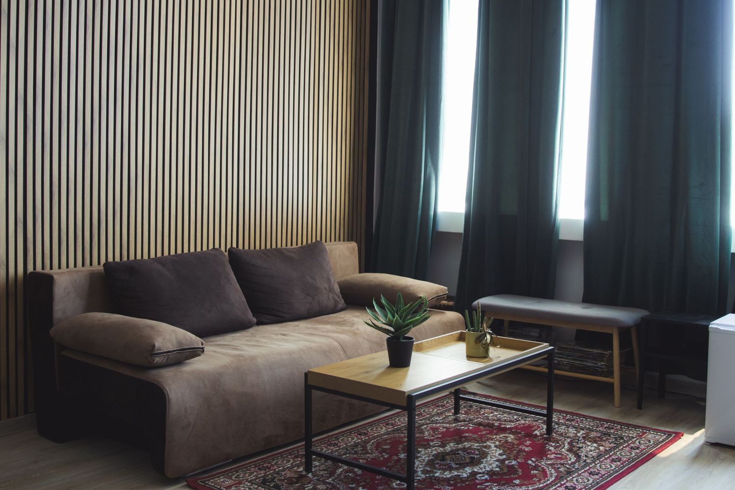 Modern living room with brown sofa, wooden slat accent wall, red patterned rug, and dark curtains.