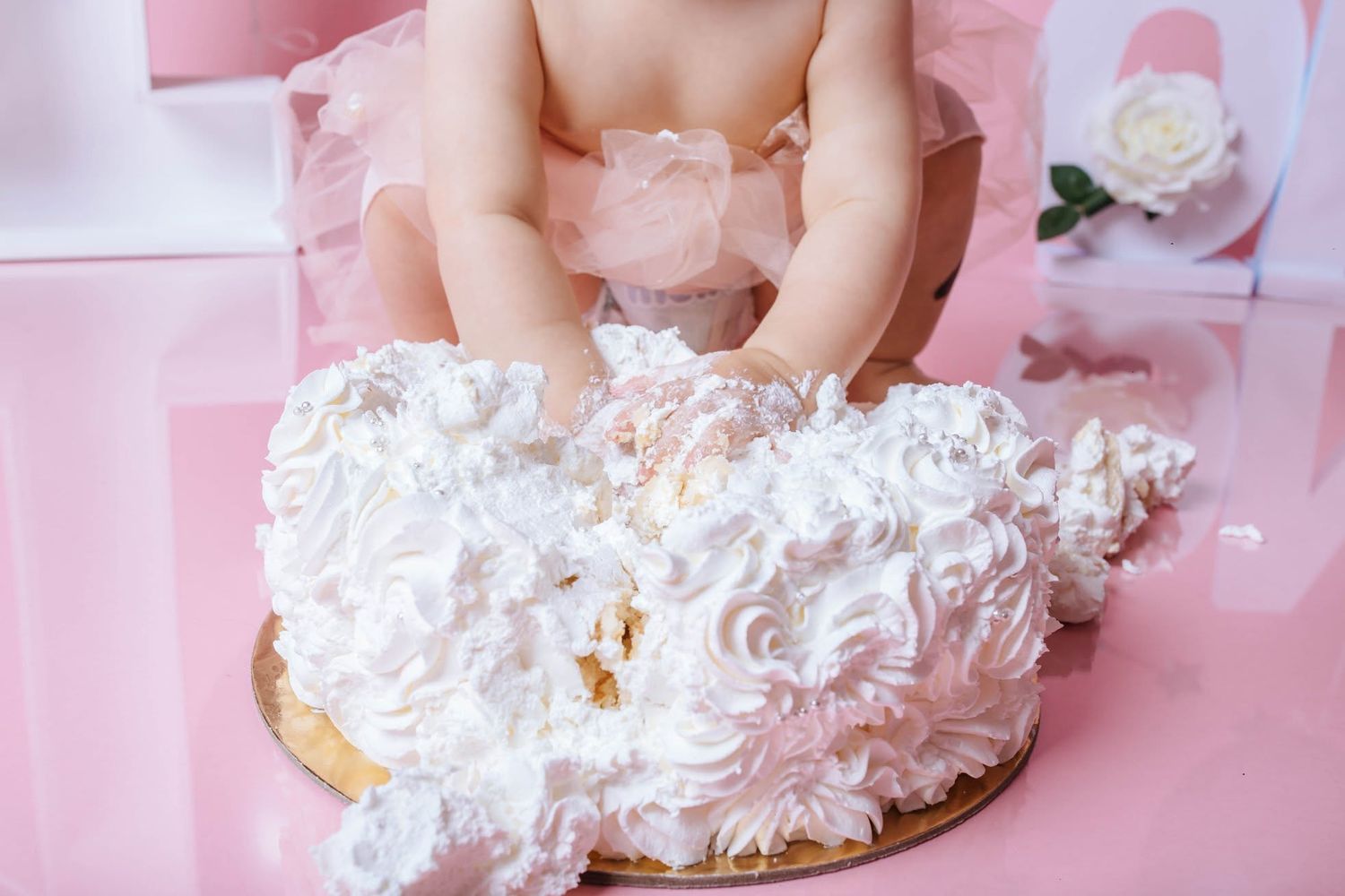 Baby in pink tutu smashing a white rosette first birthday cake on a reflective pink surface.