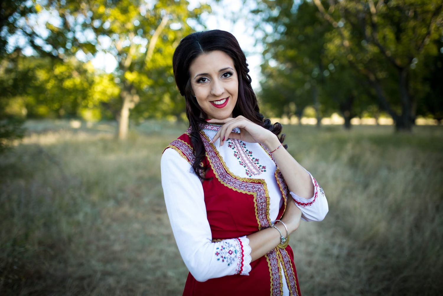A smiling woman in traditional Bulgarian folk costume with red vest and white embroidered blouse poses outdoors.