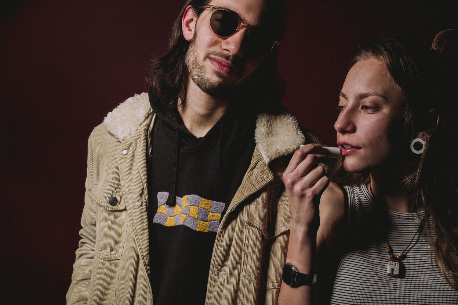 Two young adults pose together, man in sunglasses and jacket, woman smoking, against dark red background.