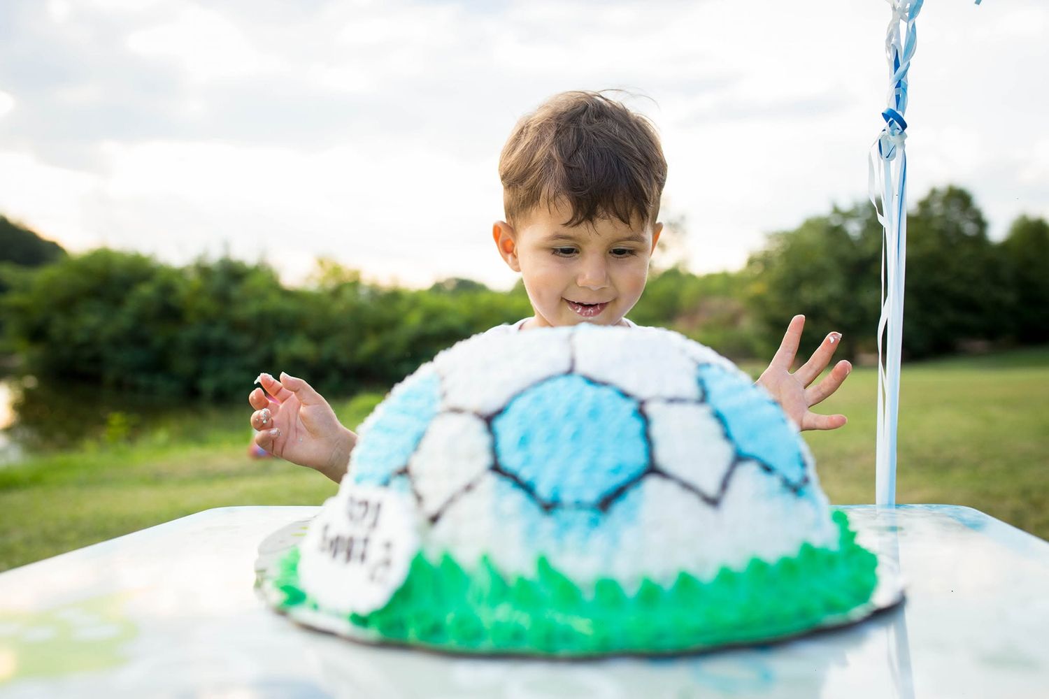 Young boy admiring a soccer ball-shaped birthday cake outdoors at a party celebration.