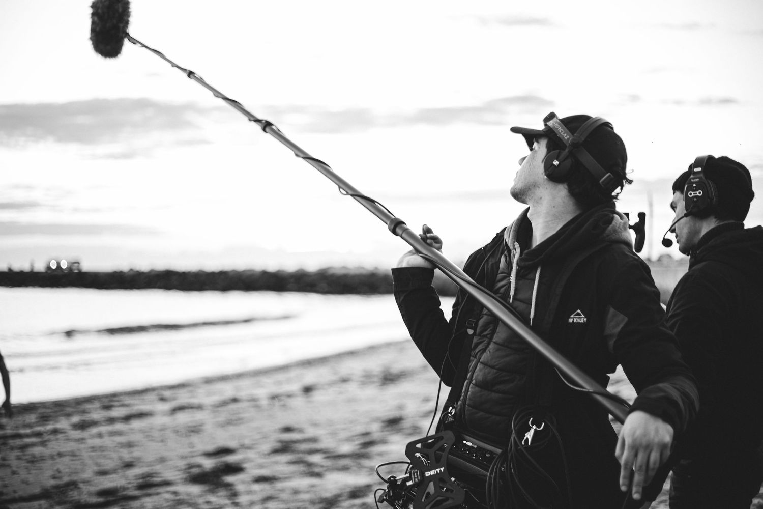 Two crew members operate a boom microphone on a beach at dusk in black and white.