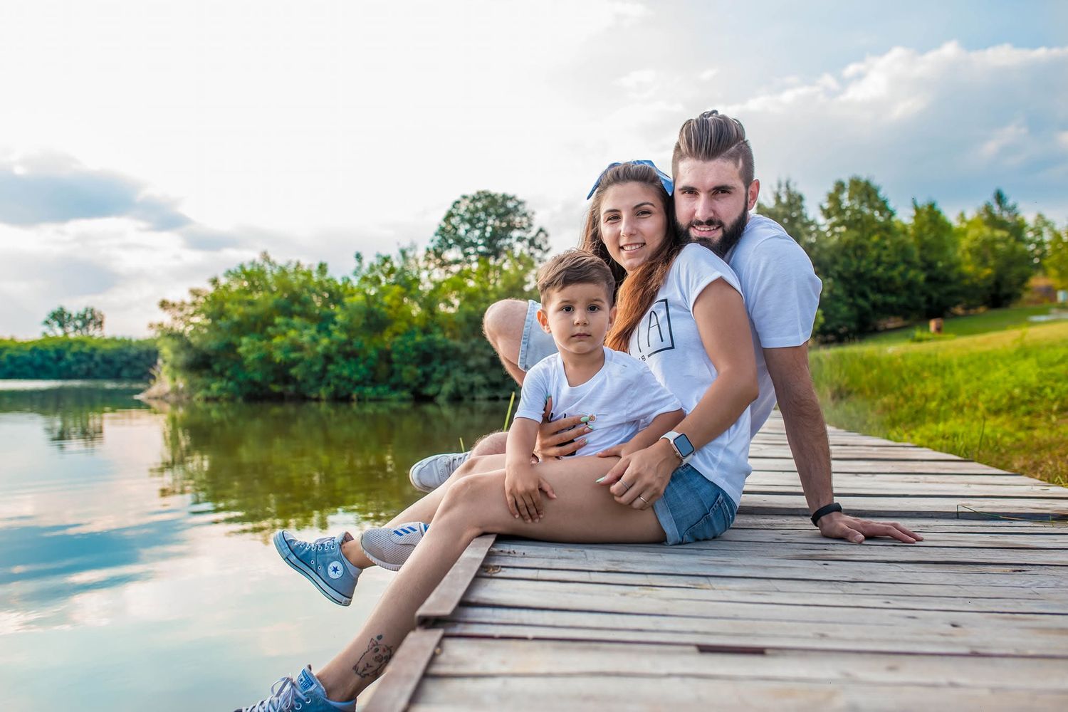 Family of three sitting on a wooden dock by a lake on a sunny day.