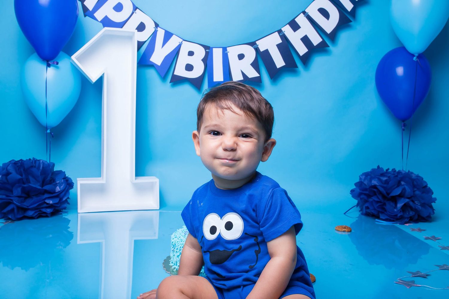 Baby boy in Cookie Monster shirt sits before a blue Happy Birthday banner and balloons celebrating his 1st birthday.
