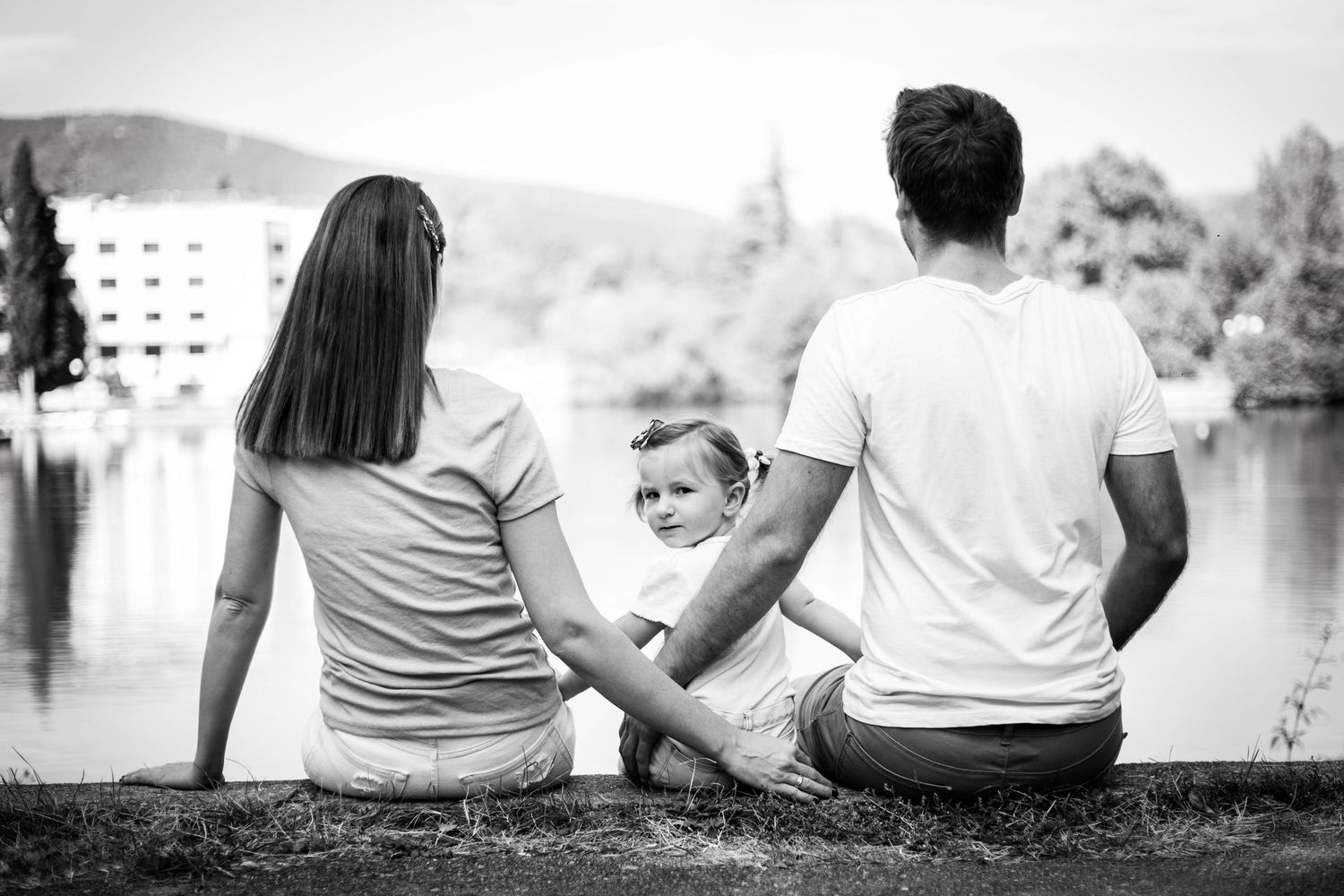 Black and white photo of couple with baby sitting by lakeside looking at scenic water view.