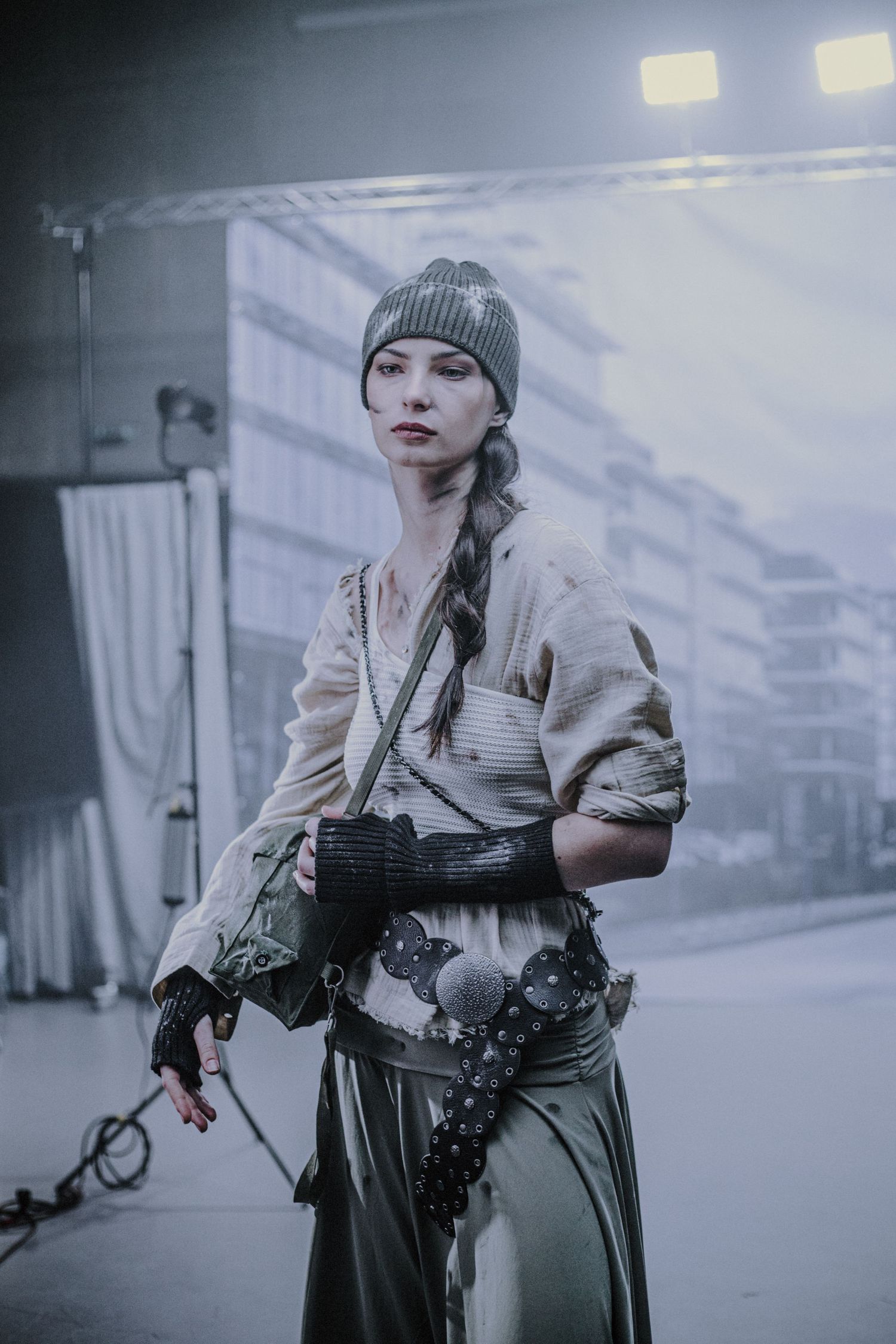 Woman in post-apocalyptic costume with beanie hat and fingerless gloves standing in foggy urban setting.