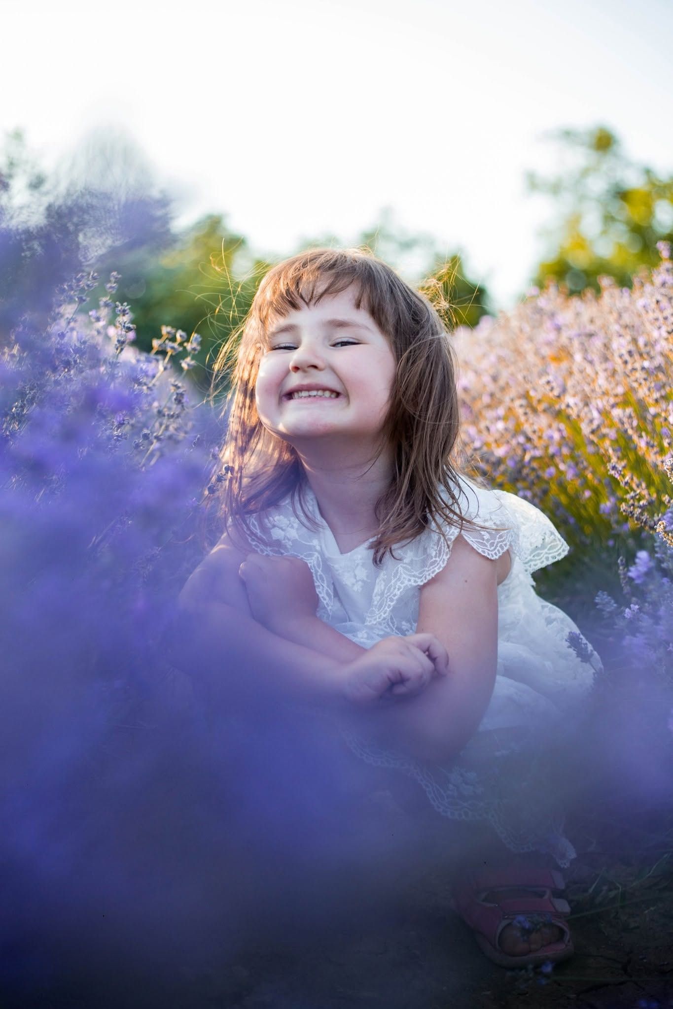 A joyful little girl in a white dress laughs with eyes closed while sitting among blooming purple lavender flowers.