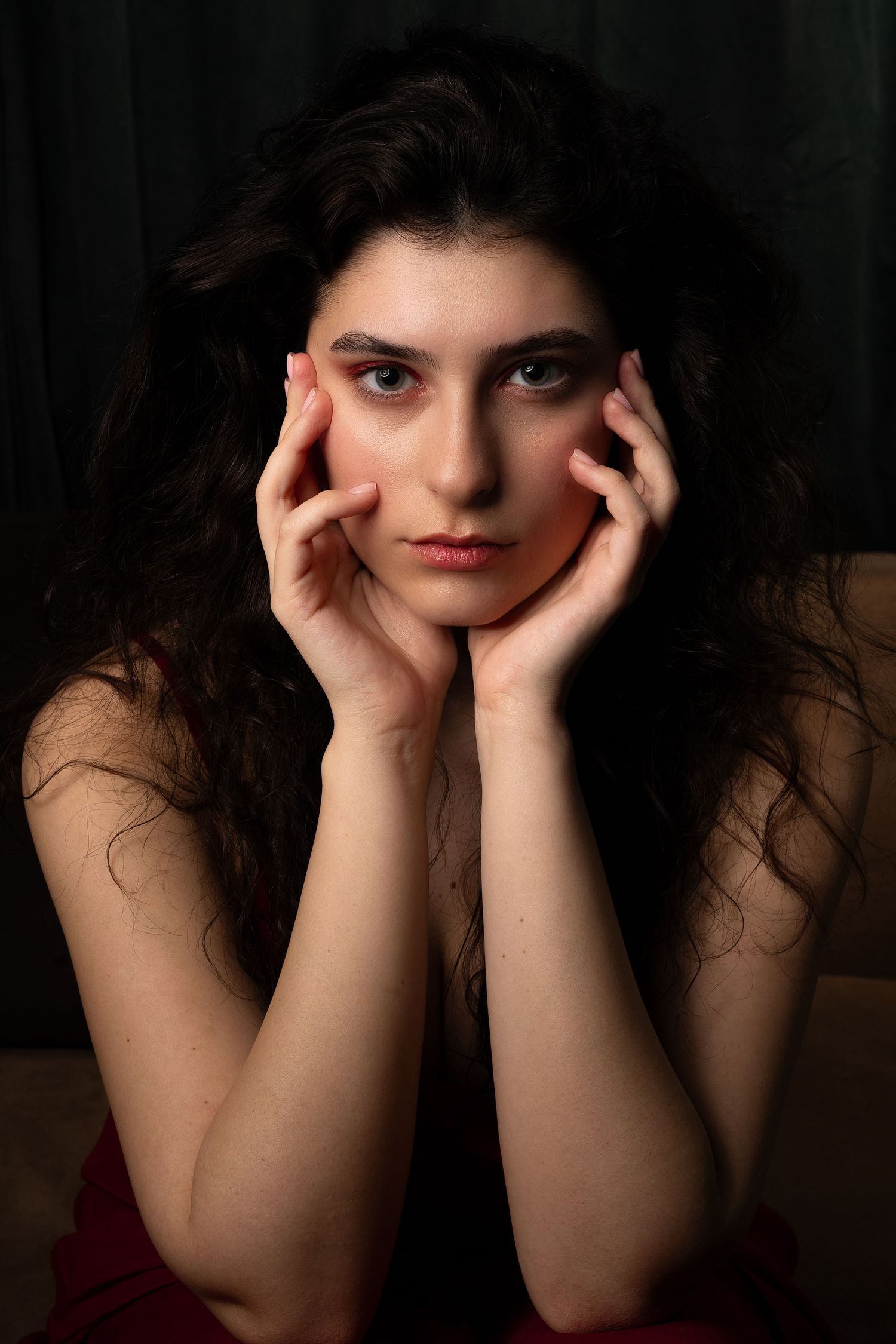 Beautiful young woman with curly dark hair holding face in hands in moody dark portrait.