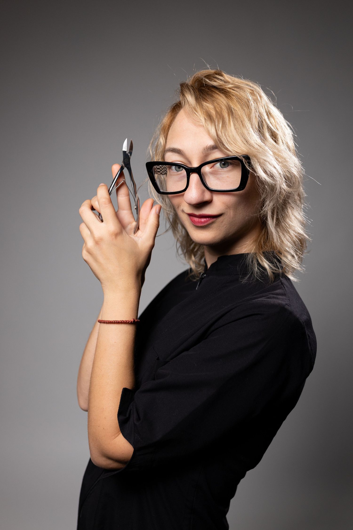 Blonde woman in black top and glasses holding nail clippers, smiling against gray background.