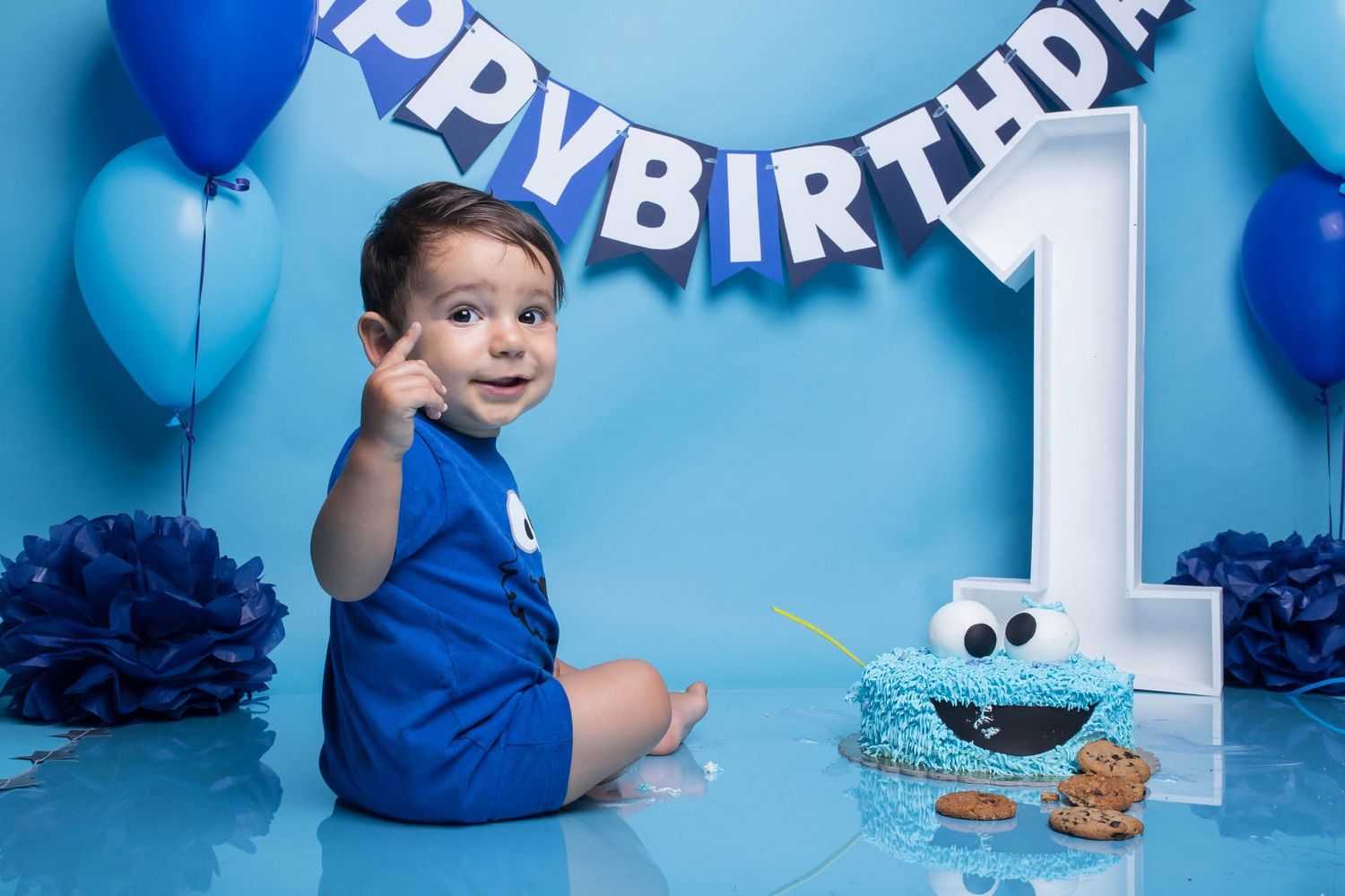 Baby boy in blue romper celebrates 1st birthday with Cookie Monster cake and blue balloons at a cake smash photo session.